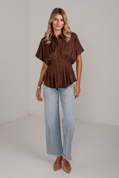 A woman modeling a brown short-sleeve button-up peplum blouse with light-wash wide-leg jeans and tan pointed-toe heels, standing against a plain white studio wall.