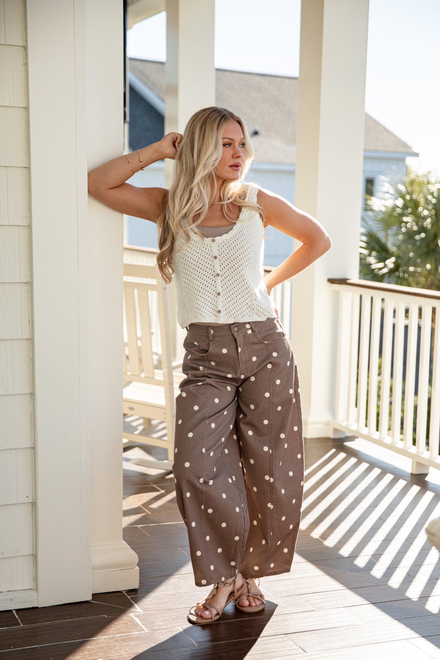 Woman standing on a porch wearing a white sleeveless top and brown polka dot pants.