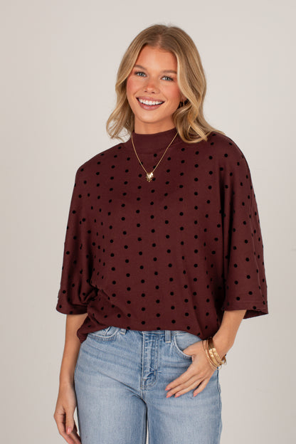 A smiling blonde woman wearing a dark brown mock-neck top with black polka dots and light-wash blue jeans, standing against a solid light gray studio background.