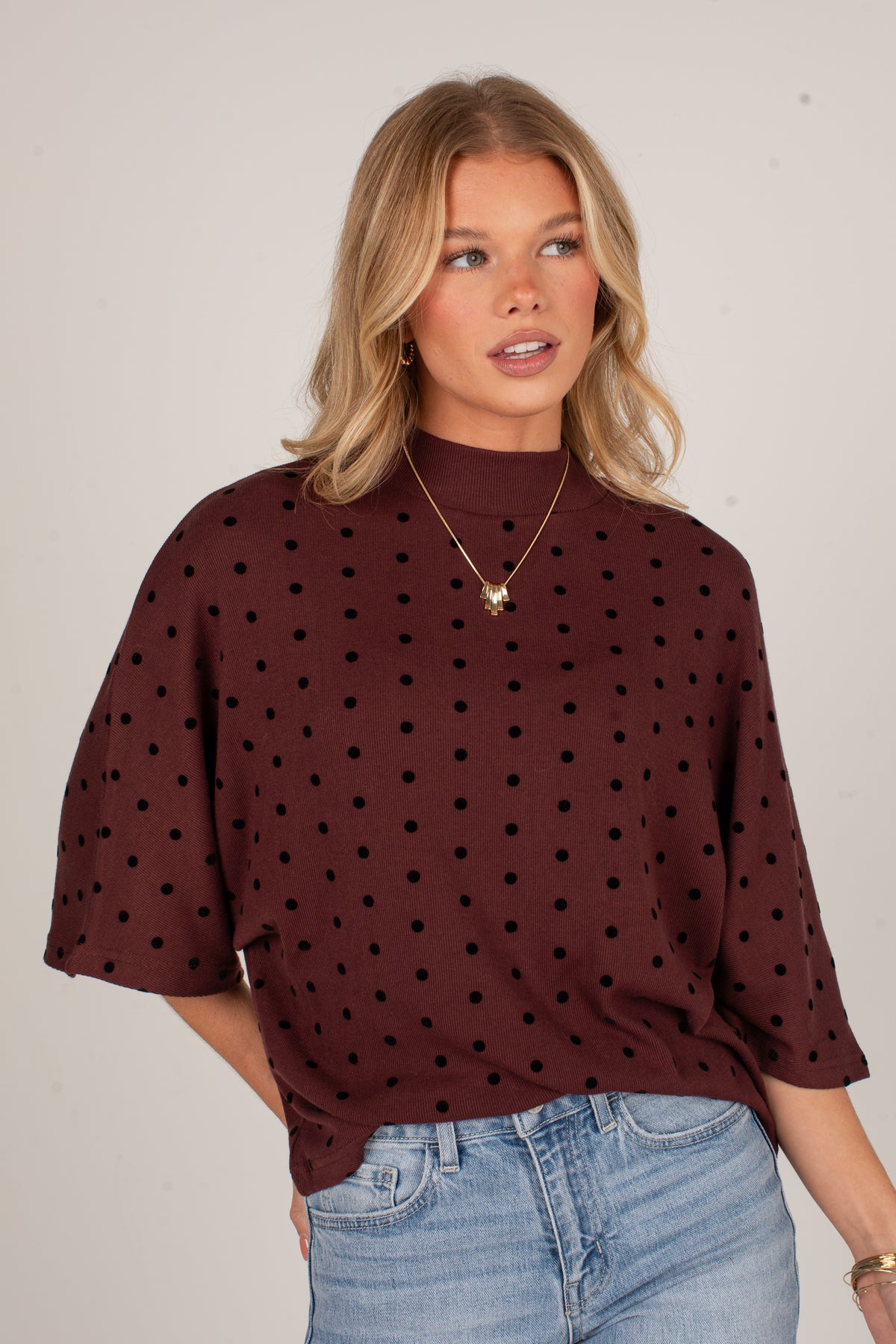 A woman wearing a dark brown mock-neck top with black polka dots and light blue jeans, standing against a plain light gray background.