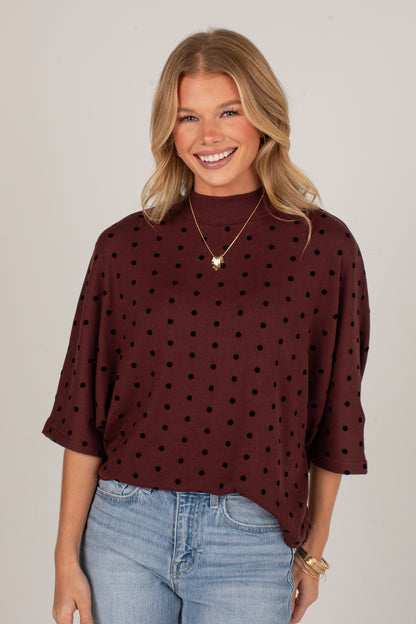 A smiling blonde woman wearing a dark brown mock neck top with black polka dots and light blue jeans, standing against a plain light gray studio background.