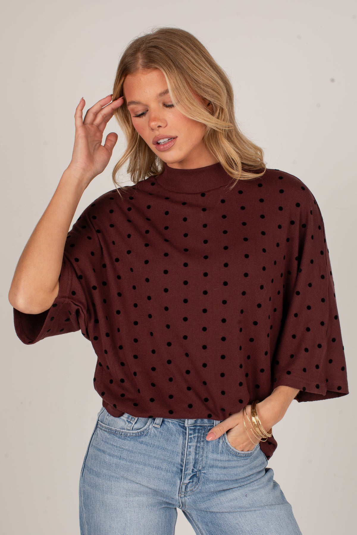 A woman wearing a dark brown mock neck top with black polka dots and wide three-quarter sleeves, paired with light blue jeans, against a plain light gray background.