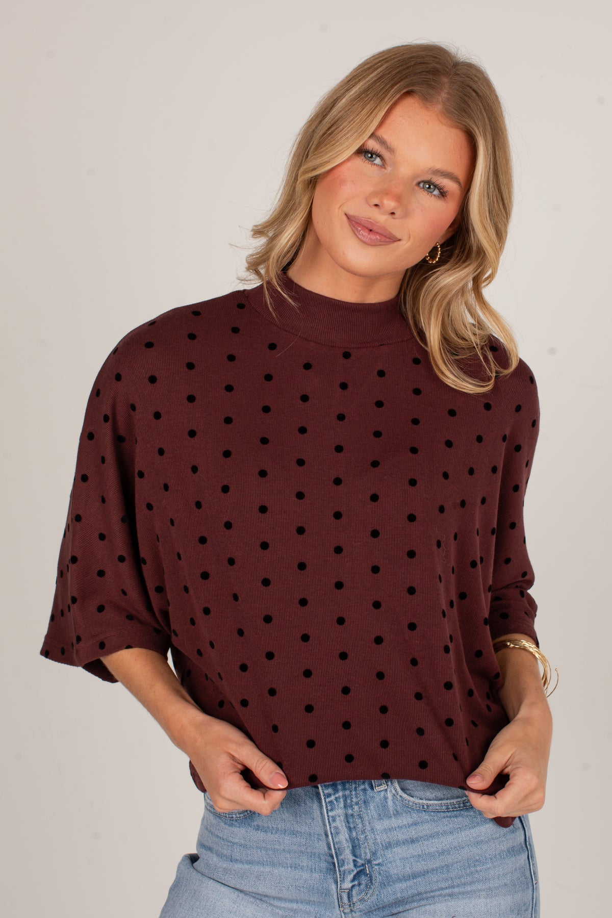 A woman wearing a dark brown mock neck top with black polka dots and light wash blue jeans, standing against a neutral light gray background.