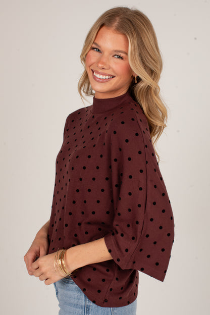 A smiling blonde woman wearing a dark brown mock neck top with black polka dots and elbow-length flared sleeves, paired with light blue jeans, photographed against a plain light gray studio background.