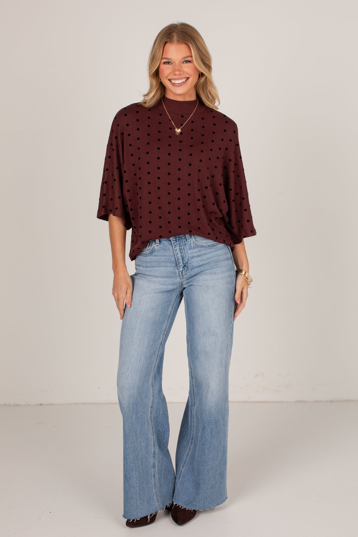 A woman modeling a dark burgundy short-sleeve top with a small black polka dot pattern, paired with light-wash blue flare jeans, standing against a plain off-white studio background.