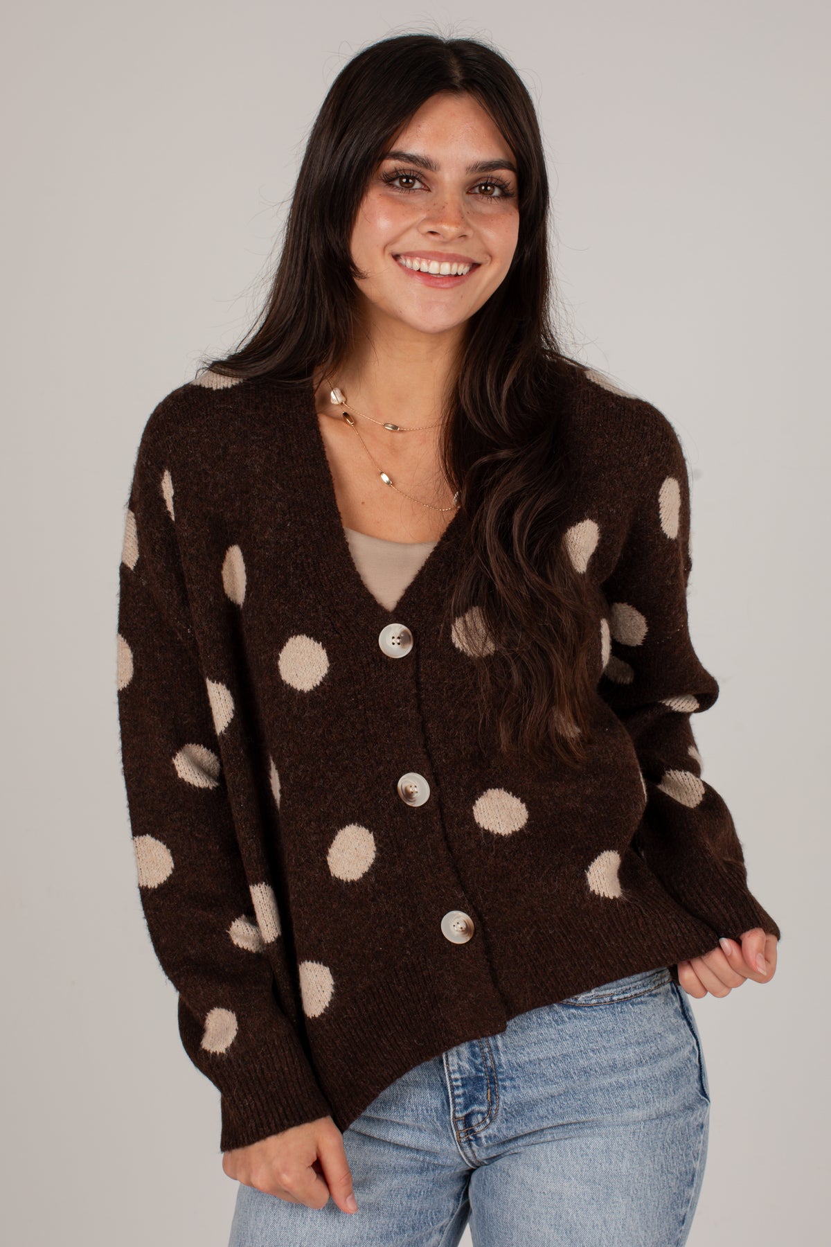 A woman wearing a dark brown knit cardigan sweater with beige polka dots and light blue jeans, standing against a plain light gray studio background.