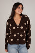 A woman wearing a dark brown knit cardigan with beige polka dots and silver buttons, paired with light blue jeans, against a plain light gray background.