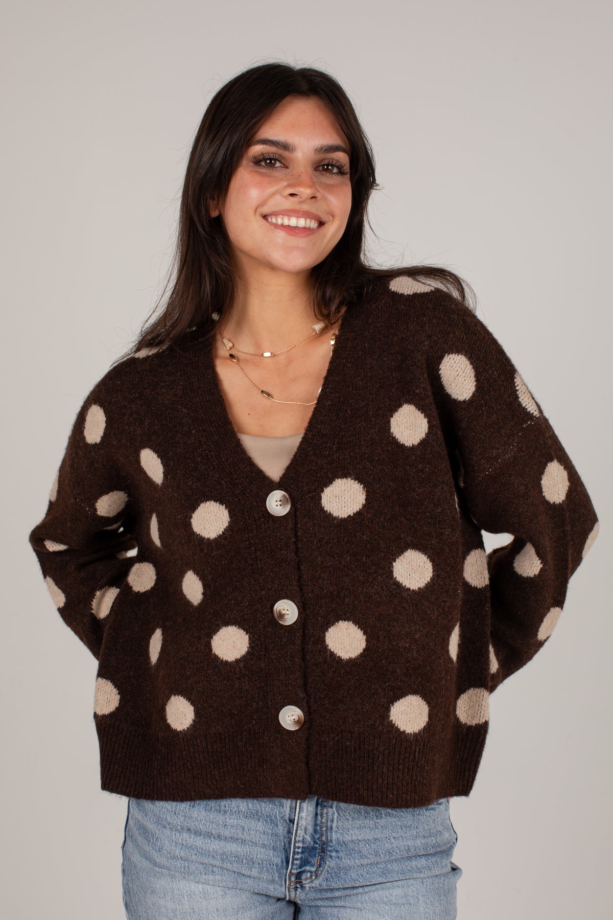 A woman wearing a dark brown knit cardigan sweater with large beige polka dots and silver buttons, paired with light blue jeans, against a plain light gray background.