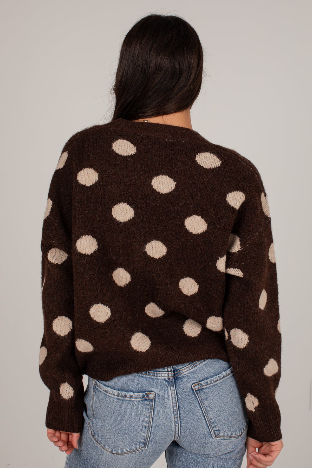 A person viewed from the back wearing a dark brown knit sweater with large beige polka dots and light blue jeans, against a plain light gray studio background.