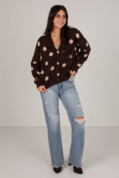 A woman modeling a dark brown button-up cardigan with large beige polka dots, paired with light-wash distressed jeans and dark brown pointed shoes, against a plain light gray studio background.