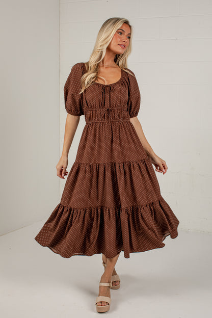A woman modeling a brown tiered midi dress with a small white polka dot pattern, puff sleeves, and a tie-neck detail, set against a white studio background.