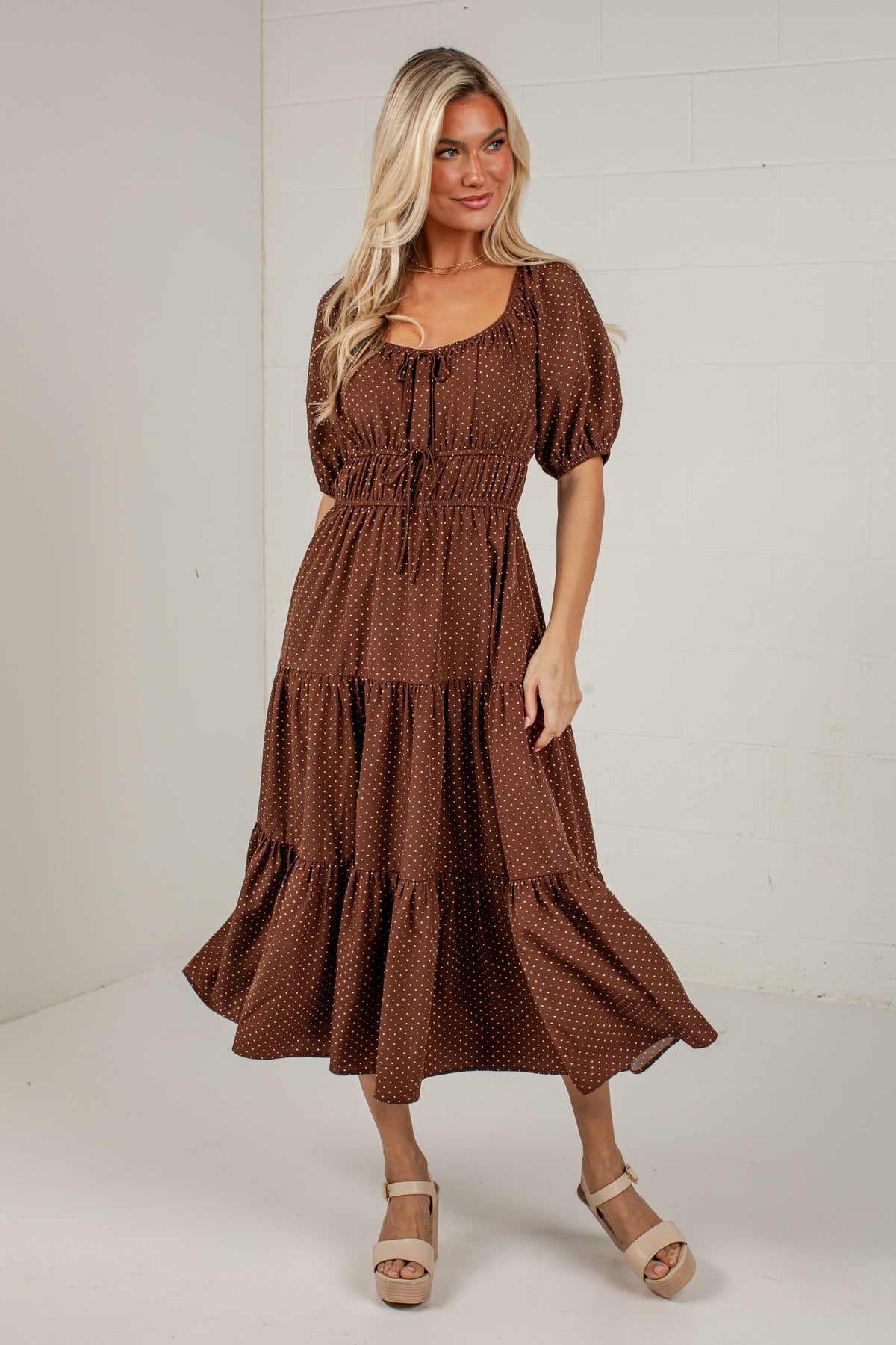 A woman modeling a brown tiered midi dress with a small white geometric pattern, puff sleeves, and a tie-neck detail, paired with beige platform sandals against a white brick wall background.