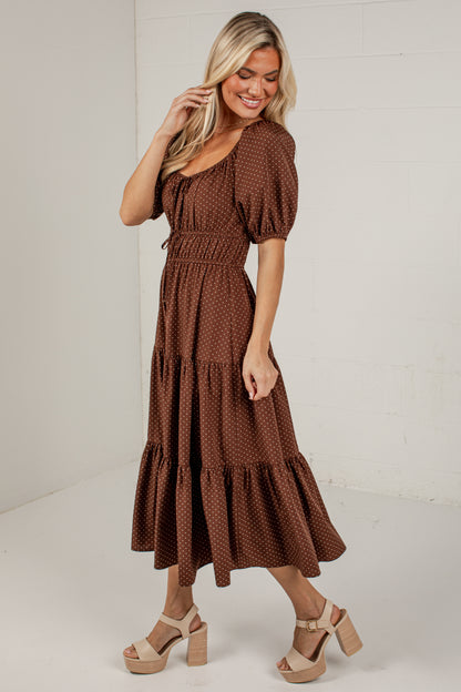 A woman modeling a brown tiered midi dress with a small white geometric pattern, puff sleeves, and a tie waist, paired with beige platform sandals in a bright studio setting.