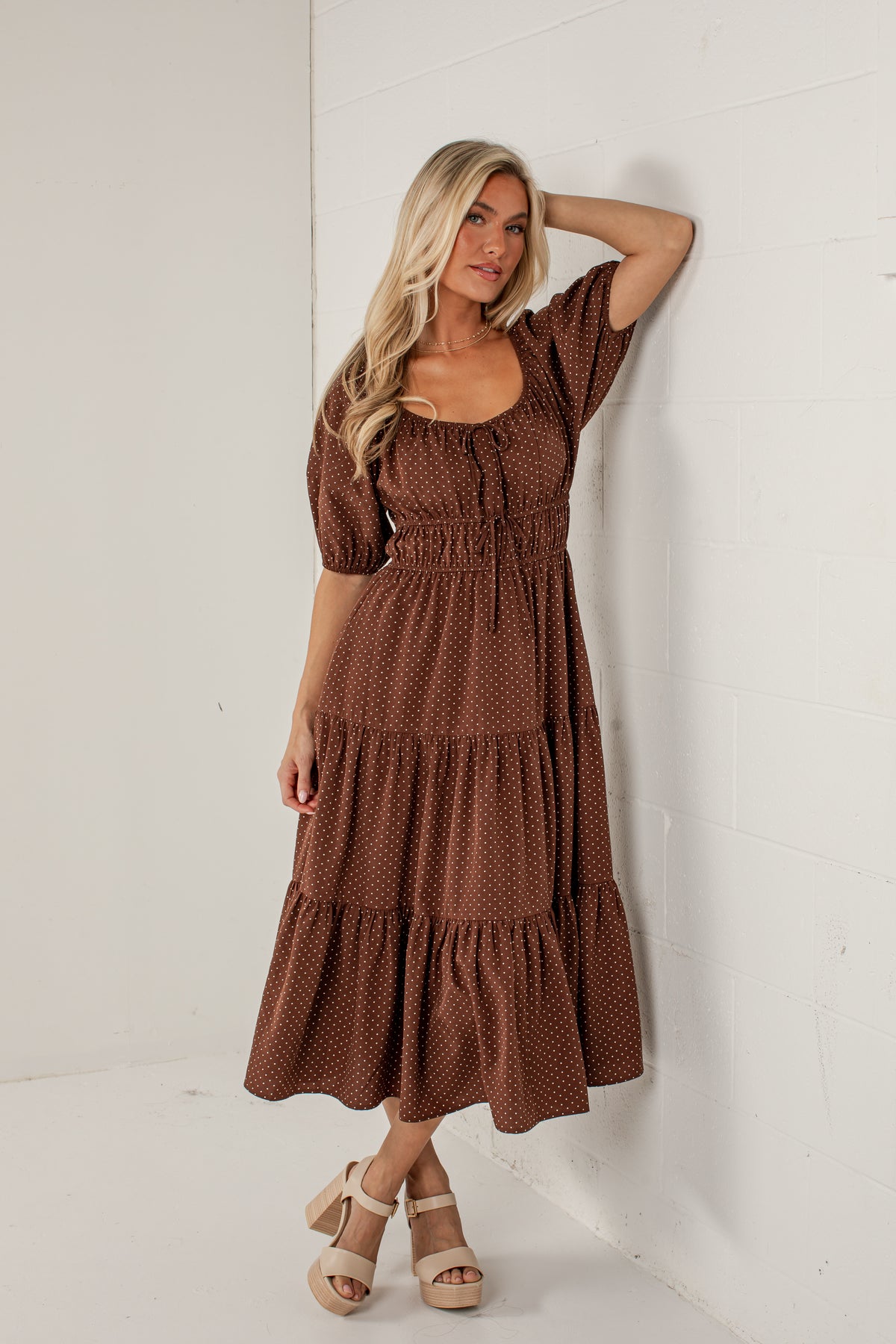 A woman modeling a brown tiered midi dress with a small white polka dot pattern, standing against a white wall.