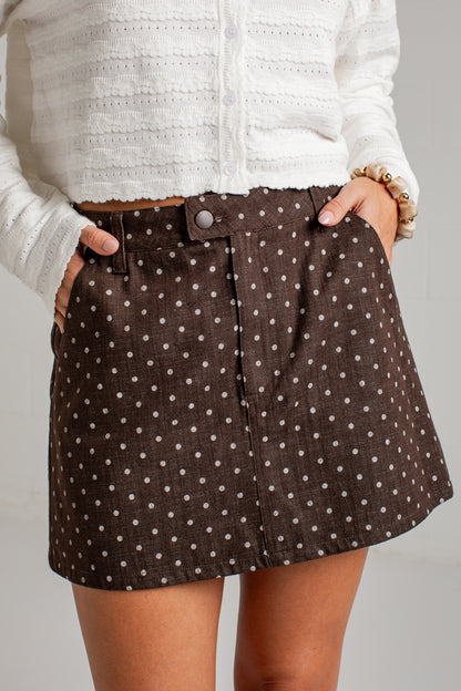 A close-up of a person wearing a brown mini skirt with a white polka dot pattern, paired with a white textured knit cardigan. The skirt features a button closure at the waistband and side pockets.