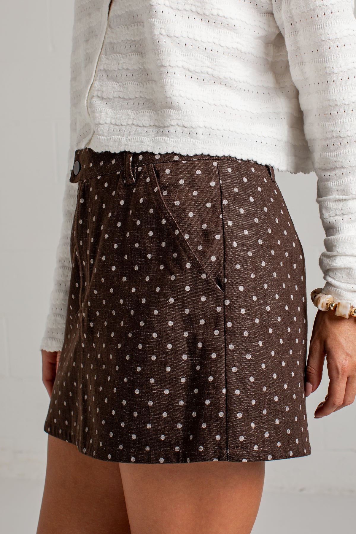 A close-up of a person wearing a brown mini skirt with a white polka dot pattern, paired with a white textured knit top.