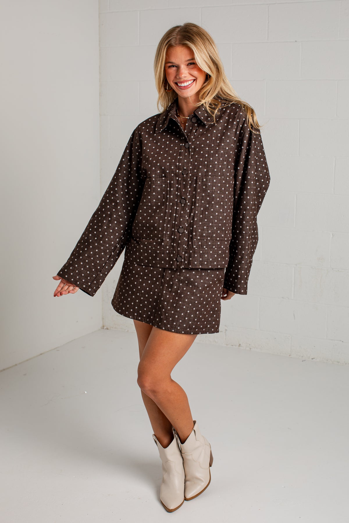 A woman modeling a brown button-up shirt dress with a small white polka dot pattern, paired with beige ankle boots, in a bright white studio setting.