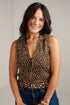 A smiling woman with dark hair wearing a brown sleeveless top with a white polka-dot pattern and tie neckline, paired with blue jeans, against a light gray background.
