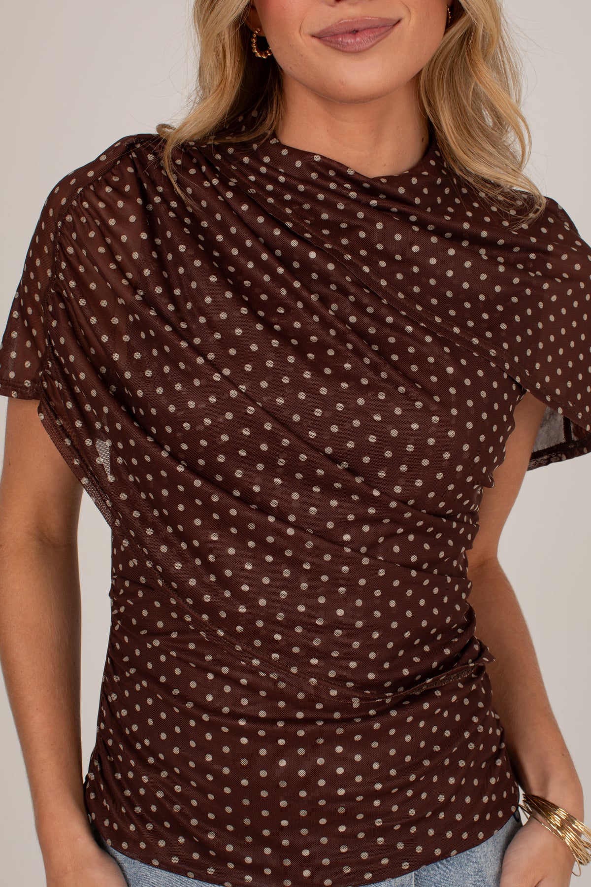A woman wearing a brown short-sleeve top with a white polka dot pattern and a draped, gathered neckline, paired with light blue jeans.