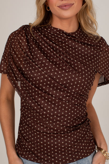 A woman wearing a brown short-sleeve top with a white polka dot pattern and a draped, gathered neckline, paired with light blue jeans.