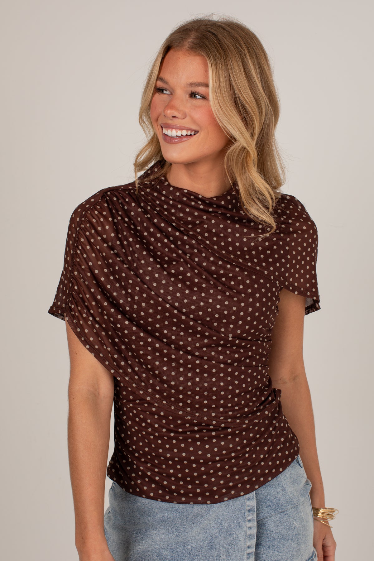 A woman wearing a brown short-sleeve top with a small white polka dot pattern, paired with light blue jeans, against a plain light gray background.