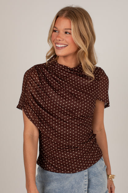 A woman wearing a brown short-sleeve top with a small white polka dot pattern, paired with light blue jeans, against a plain light gray background.