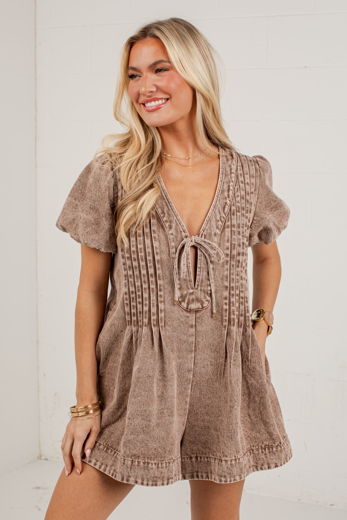 A woman modeling a short-sleeve romper in a textured taupe-brown fabric with a tie-front detail and vertical stitched accents, posed against a white brick wall.