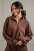 A woman wearing a brown half-zip sweatshirt with a drawstring waist, photographed against a light neutral wall.
