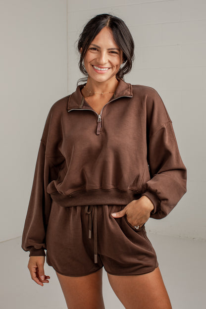 A woman modeling a matching brown lounge set consisting of a half-zip sweatshirt and drawstring shorts, photographed against a light gray studio wall.