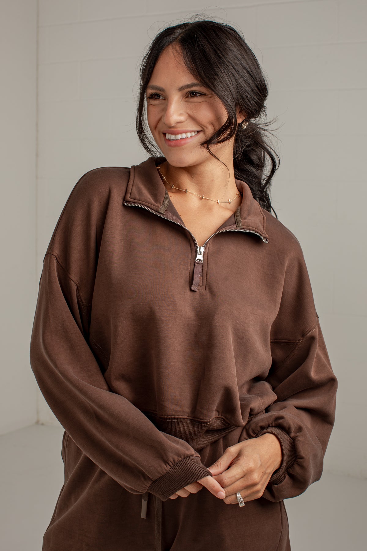 A woman wearing a brown long-sleeve quarter-zip sweatshirt with a drawstring waist, standing against a light gray wall.
