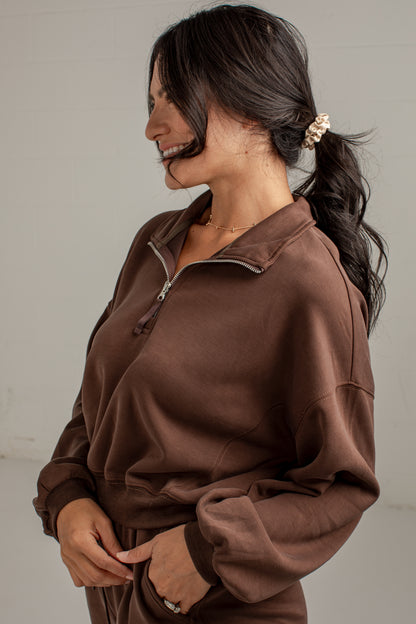 A woman wearing a brown long-sleeve quarter-zip sweatshirt, shown from a side profile against a plain light gray background.