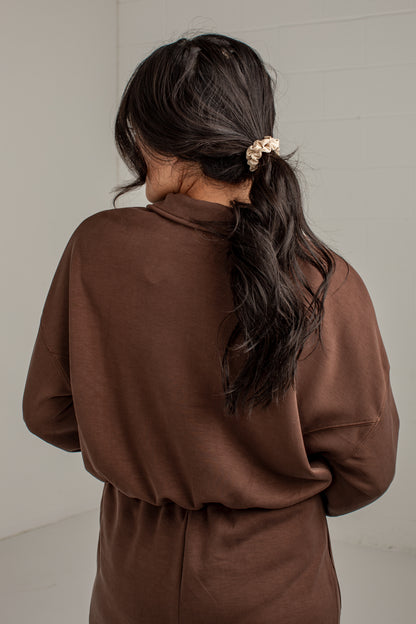 A woman viewed from behind wearing a brown long-sleeve top or dress, with her dark hair tied back in a low ponytail using a light scrunchie, against a plain light gray wall.