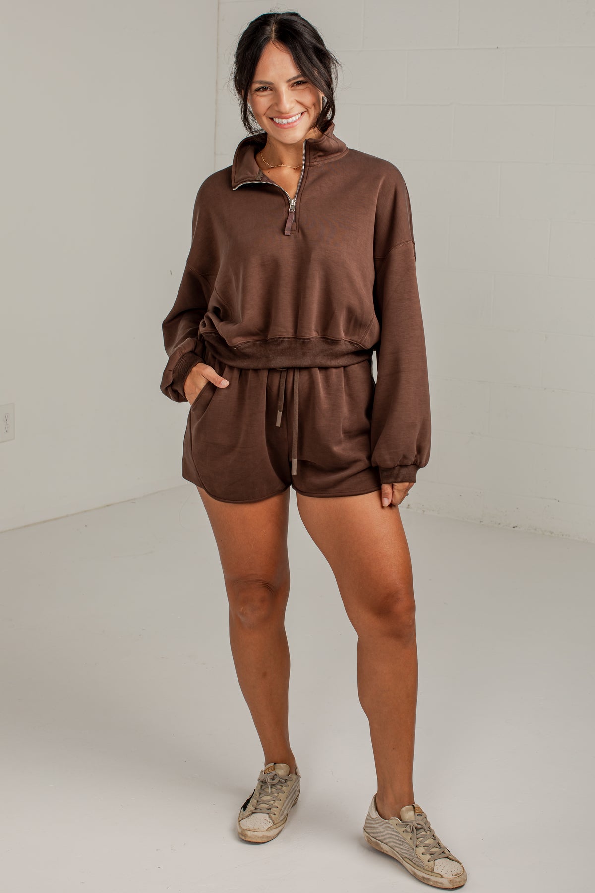 A woman modeling a matching brown lounge set consisting of a quarter-zip sweatshirt and drawstring shorts, paired with beige sneakers, in a bright studio setting.