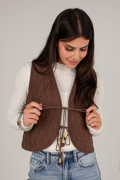 A woman wearing a brown diamond-quilted sleeveless vest over a white long-sleeve top and blue jeans, holding the front tie closure of the vest against a plain light gray background.