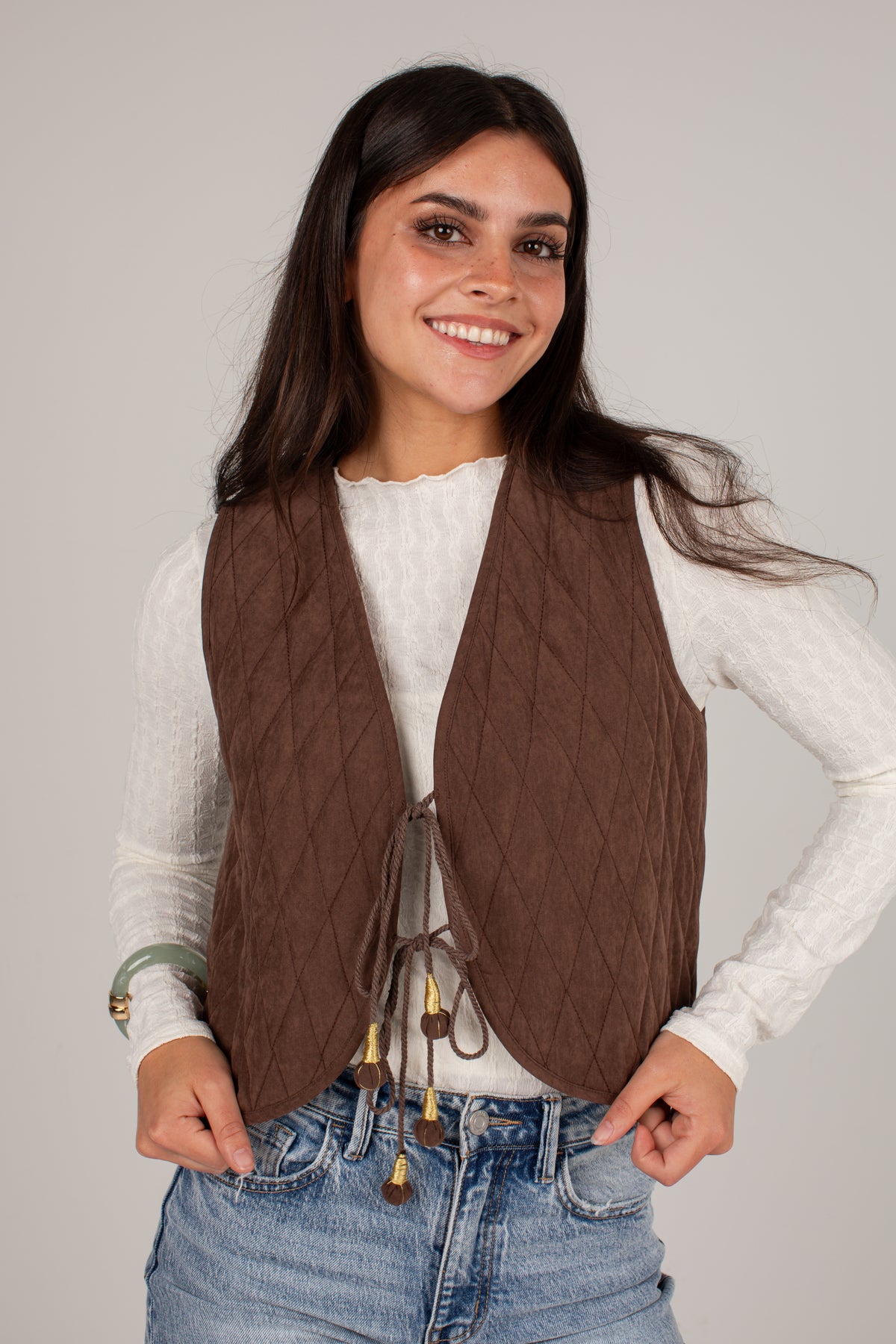 A woman wearing a brown diamond-quilted sleeveless vest over a white long-sleeve top and blue jeans, standing against a light gray studio background.