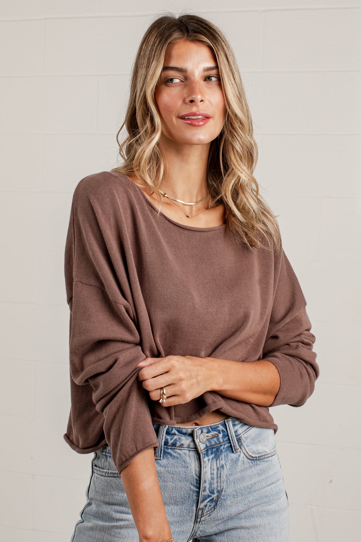 A woman wearing a brown long-sleeve knit top and light-wash blue jeans, standing against a white paneled wall.