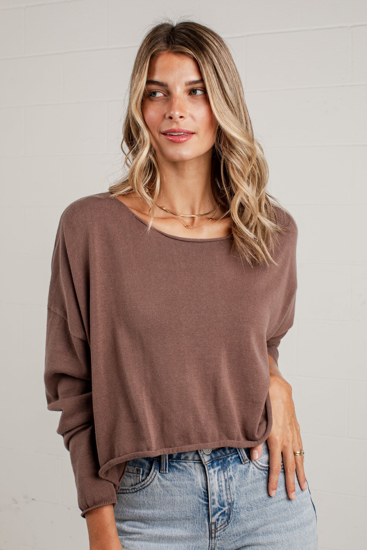 A woman wearing a brown long-sleeve knit top and light blue jeans, standing against a white brick wall.