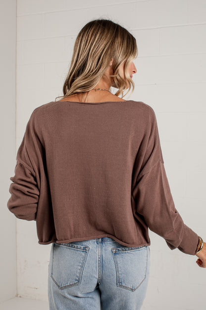 A woman viewed from behind wearing a brown long-sleeve knit top and light blue jeans, standing against a white paneled wall.
