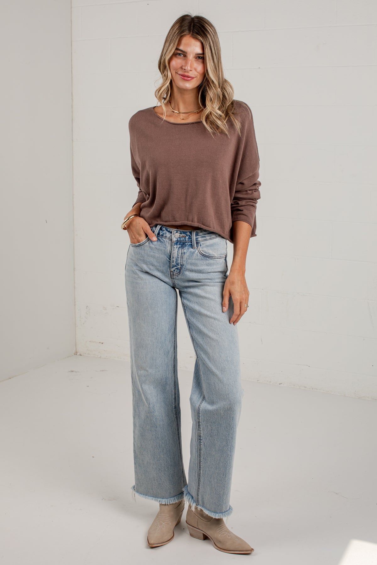 A woman modeling a brown long-sleeve knit top and light-wash wide-leg jeans with beige ankle boots, standing against a white studio wall.