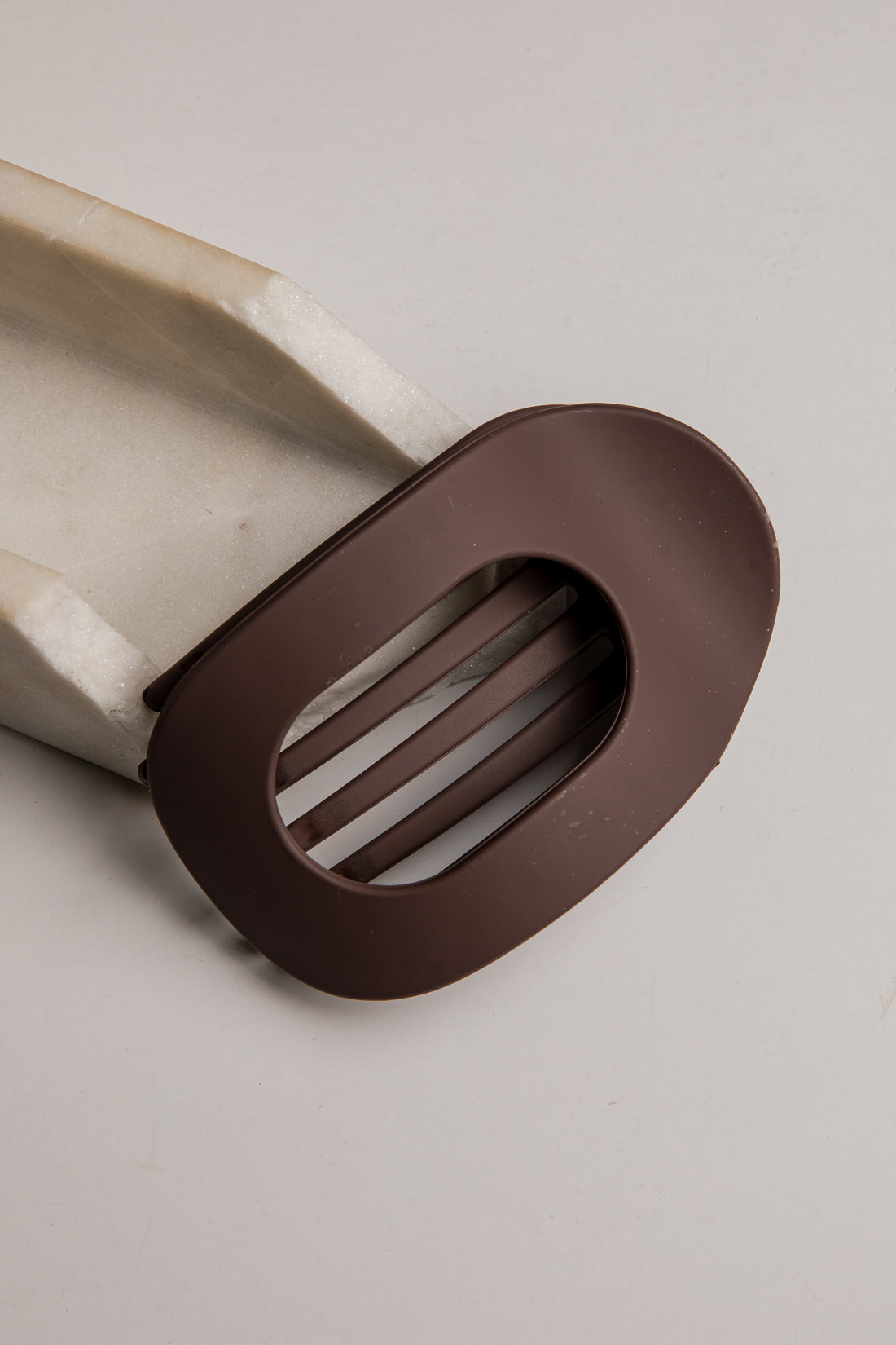 A close-up of a dark brown silicone or rubber kitchen utensil component with a slotted oval cutout, attached to a light beige handle, on a neutral light gray background.