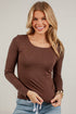 A smiling blonde woman wearing a fitted long-sleeve scoop-neck top in dark brown, paired with light blue jeans, standing against a white paneled wall.