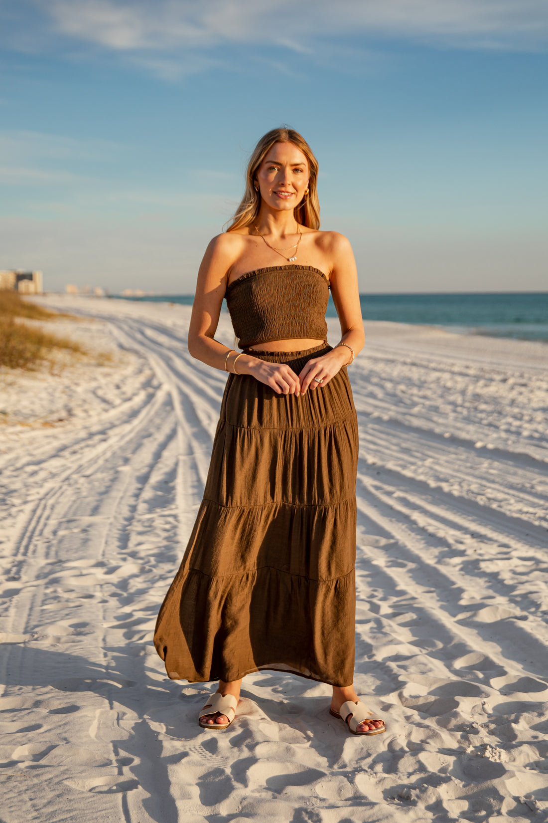 Woman in a brown dress standing on a sandy beach with ocean and sky in the background