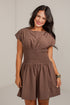 A woman modeling a brown short-sleeve dress with a smocked waist and flared skirt, standing against a light gray wall.