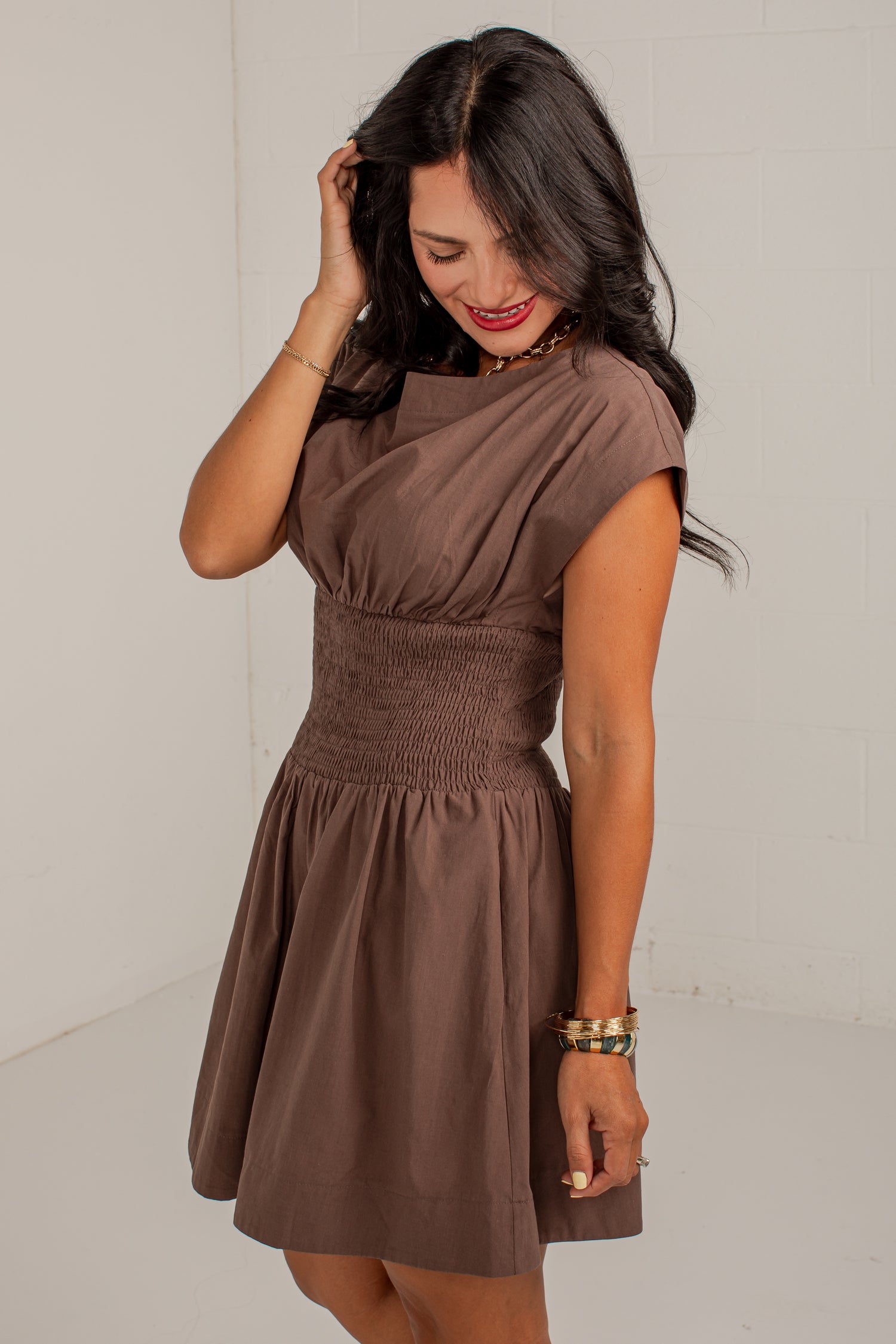 A woman wearing a brown short-sleeve dress with a gathered smocked bodice and flared skirt, standing against a white wall.