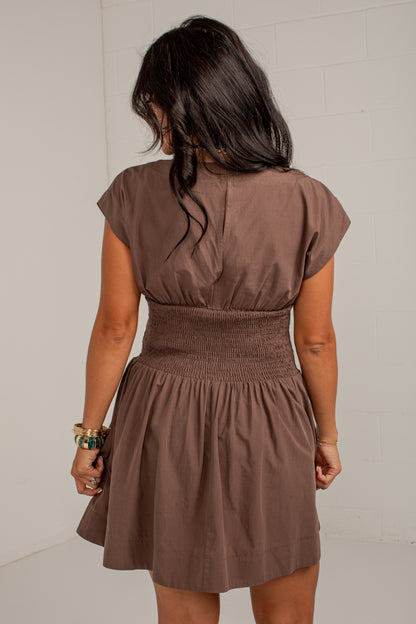 A woman viewed from behind wearing a brown short-sleeve dress with a smocked waist and flared skirt, standing against a light neutral wall.