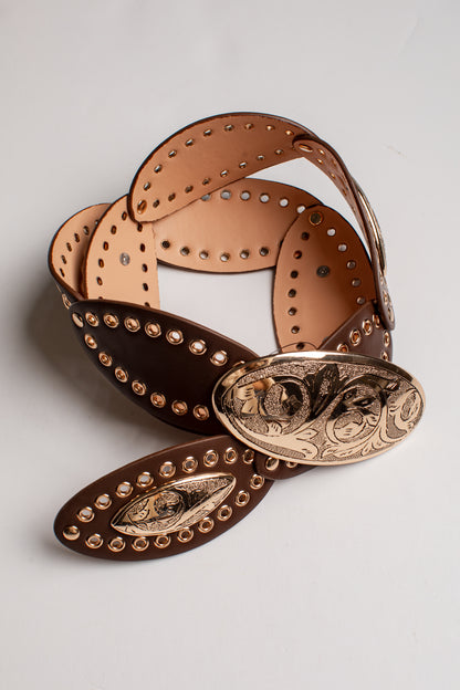 A brown leather belt with a large engraved gold-tone oval buckle, studded along the edges, coiled on a light gray background.