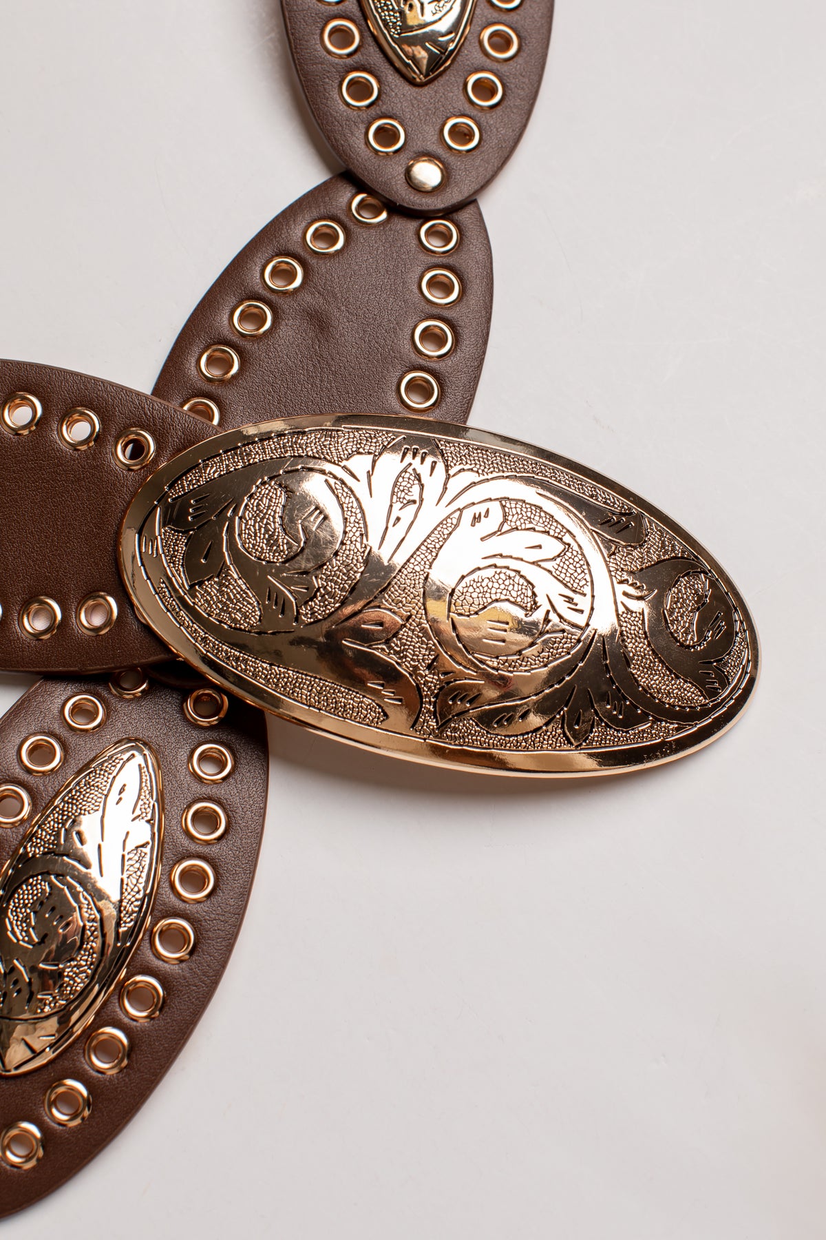 A close-up of a brown leather belt with a large, ornate oval buckle in a warm gold/bronze tone, decorated with floral engravings and surrounded by rows of gold studs.