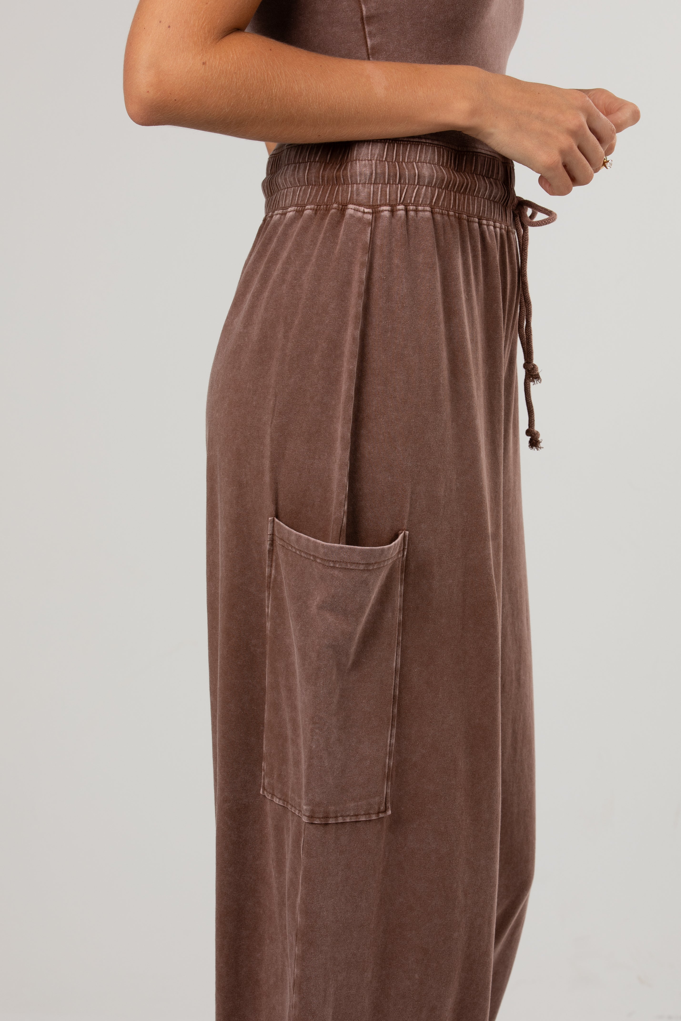 A close-up of a person wearing loose, wide-leg brown corduroy pants with an elastic waistband, drawstring, and a large front patch pocket, against a plain light gray studio background.