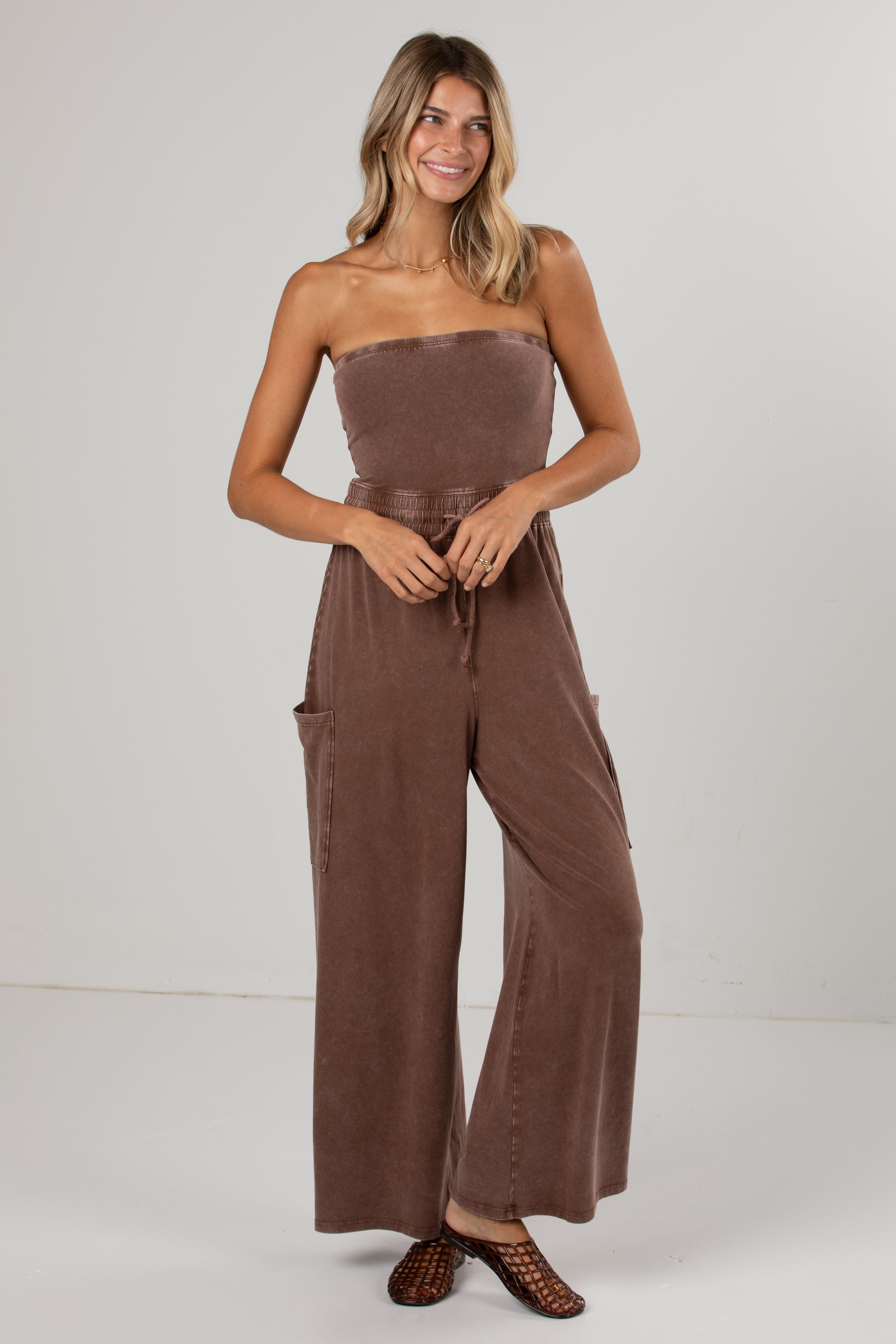 A woman modeling a brown strapless wide-leg jumpsuit with a matching tie belt at the waist, photographed against a plain light gray studio background.