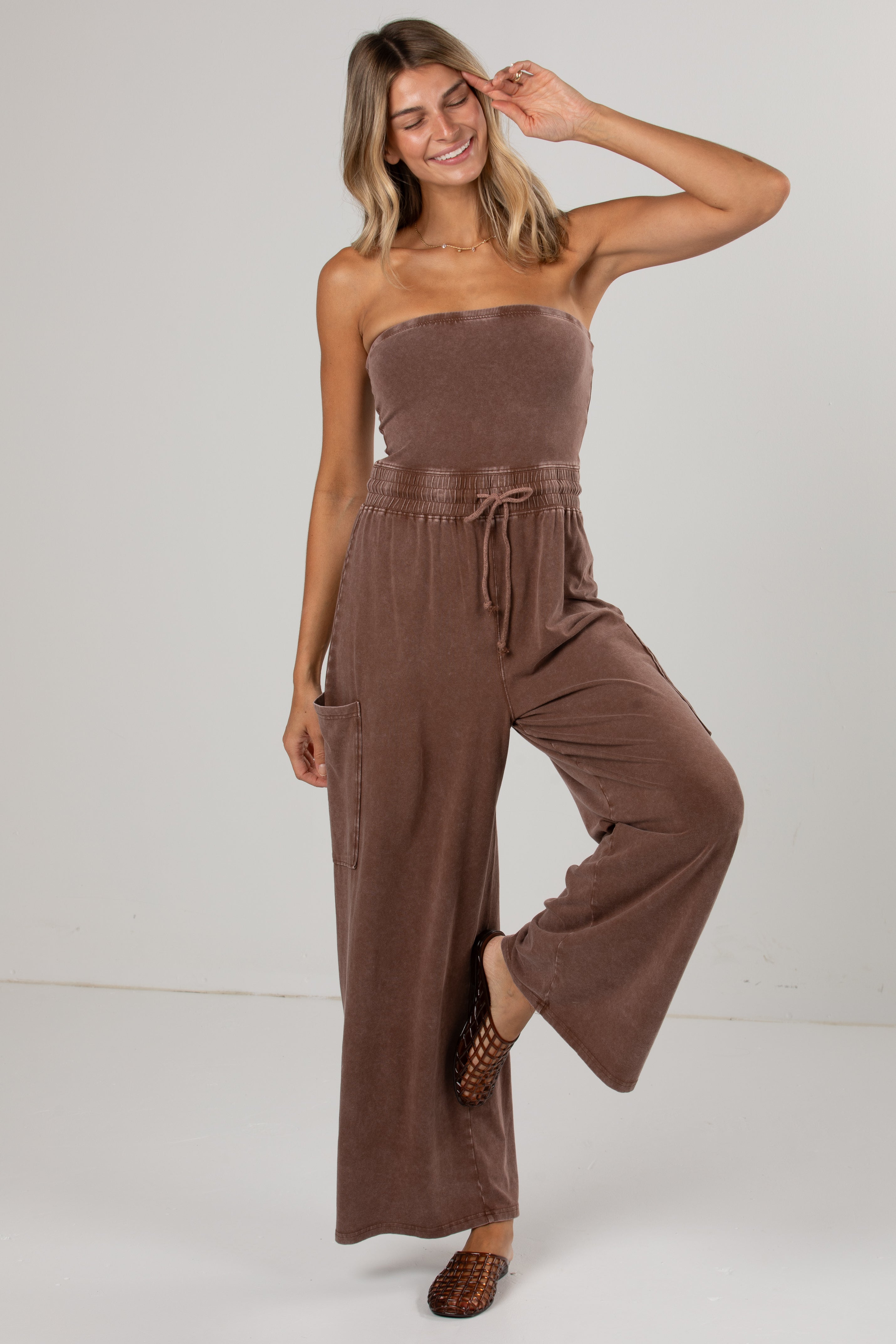 A woman modeling a brown strapless jumpsuit with wide-leg pants and a drawstring waist, photographed against a plain light gray studio background.